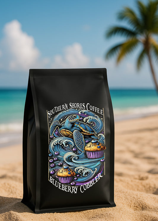 A photo of a blueberry coffee bag sitting in the sand on the beach, the ocean and a palm tree is in the background.