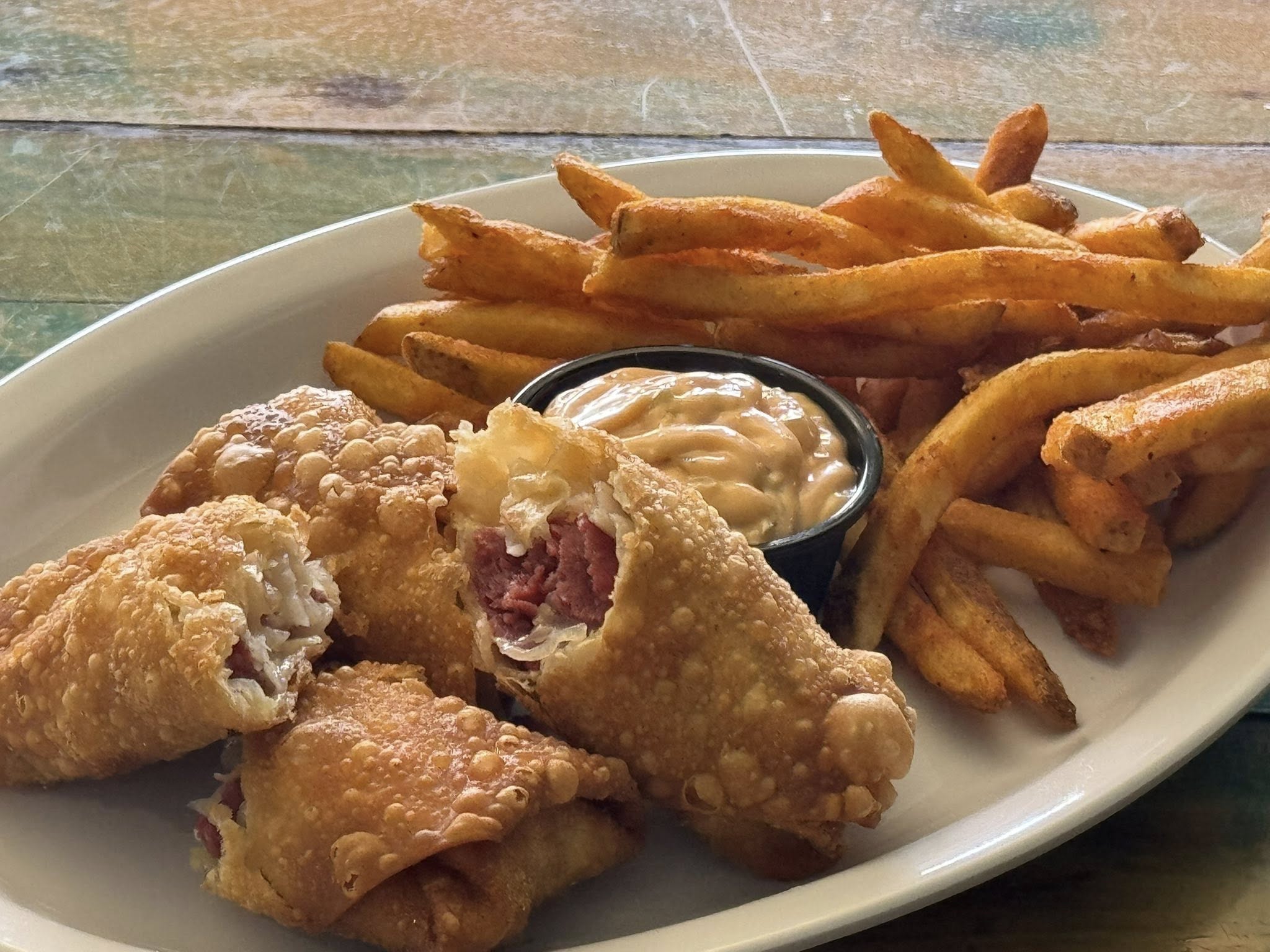 Platter of fried reuben egg rolls and French fries with a side of dipping sauce on a wooden table.